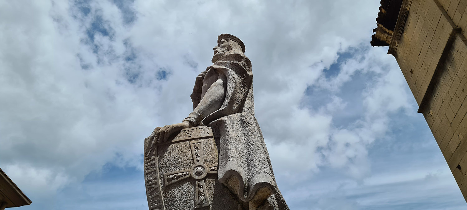 Foto en contrapicado de la escultura de Alfonso II 'El Casto' realizada en piedra. Se representa al monarca de pie, sosteniendo en la mano izquierda un escudo con el lema de la monarquía ovetense: 'Hoc signo tuetur pius. Hoc signo vincitur inimicus' y la Cruz de los Ángeles.