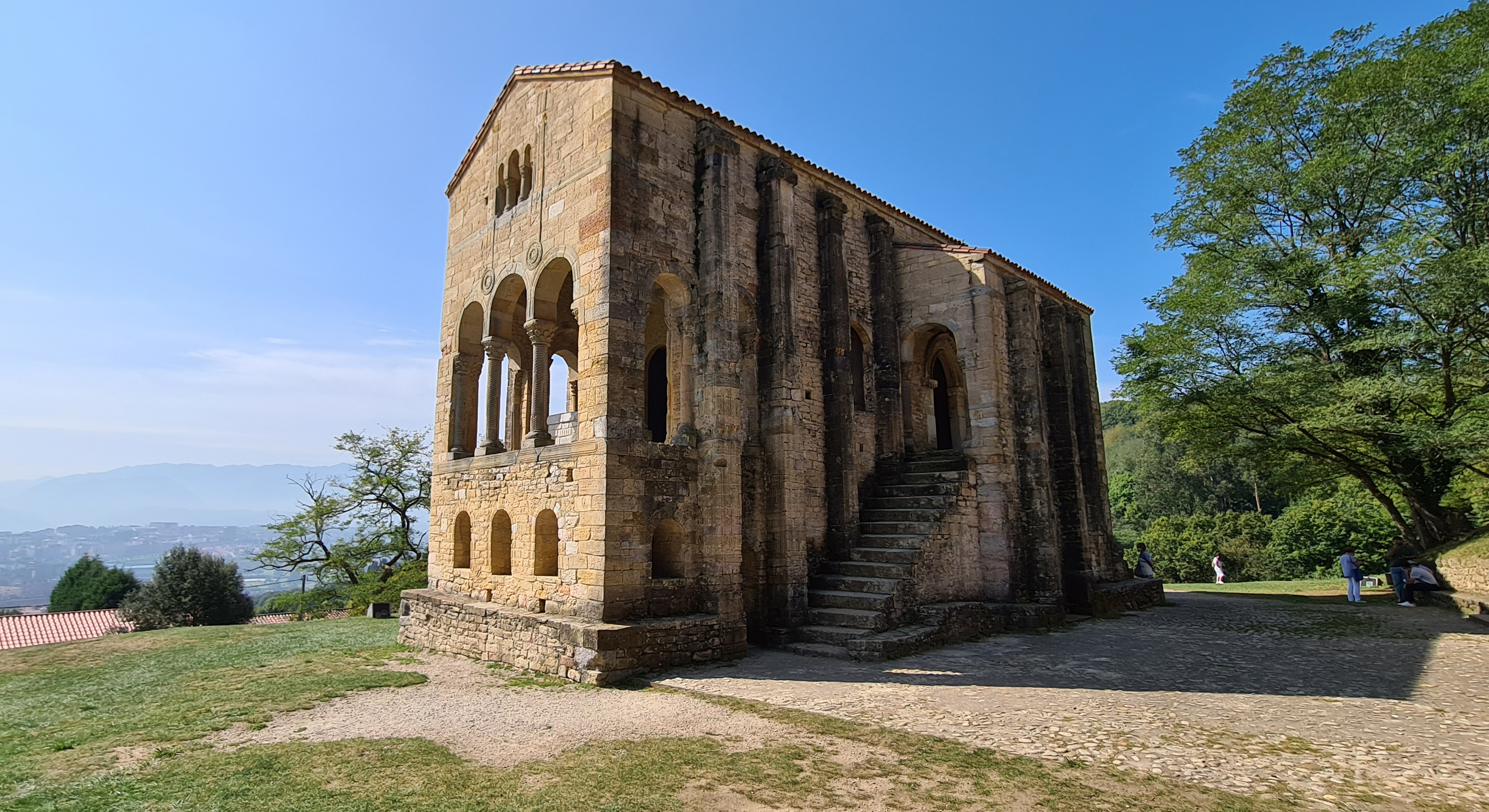 Fotografía de la iglesia prerrománica Santa María del Naranco  situada en la ladera del monte Naranco, se observa su planta rectangular con dos pisos de altura destacando al exterior los contrafuertes y la fachada con un mirador que se abre al exterior mediante tres arcos siendo el central ligeramente mayor, además de una escalera de acceso a la cámara del tesoro noble.