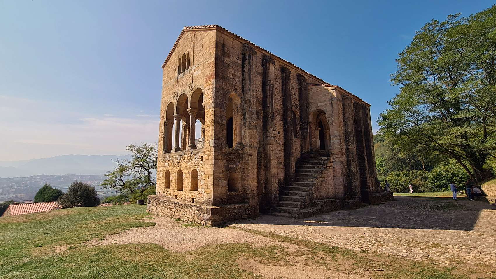 Fotografía de la iglesia prerrománica Santa María del Naranco  situada en la ladera del monte Naranco, se observa su planta rectangular con dos pisos de altura destacando al exterior los contrafuertes y la fachada con un mirador que se abre al exterior mediante tres arcos siendo el central ligeramente mayor, además de una escalera de acceso a la cámara del tesoro noble.